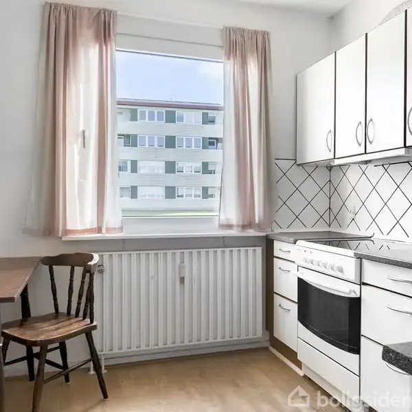 Et lyst køkken med hvide skabe og sort bordplade. Der står en stol og et bord ved vinduet, hvor man ser en bygning udenfor. Gardinerne er lyserøde.