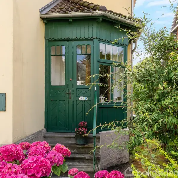 Grøn hoveddør med glaspartier, omkranset af en frodig have med lyserøde hortensiaer. Skilt på muren viser "24 December". Træ og buske kompletterer den idylliske indgang.