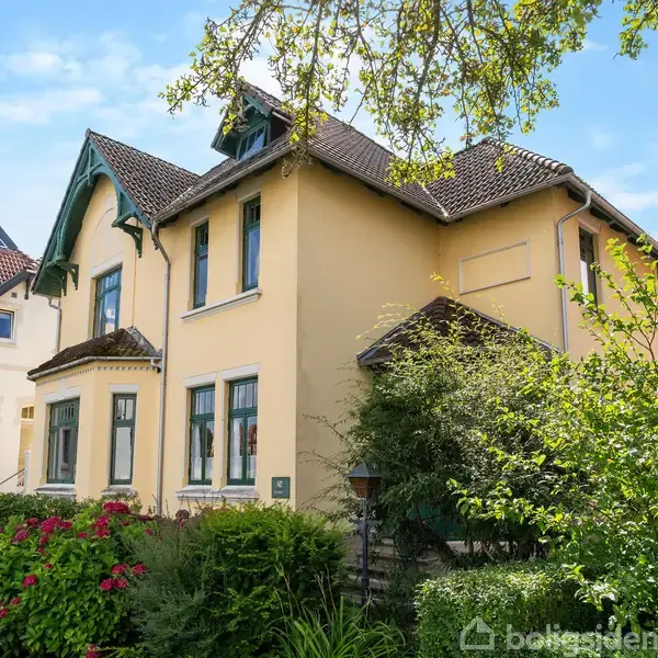 En gul to-etagers villa står i en have med grønne buske og blomster; huset har brune tage og vinduesrammer, omgivet af andre huse og træer under en blå himmel.