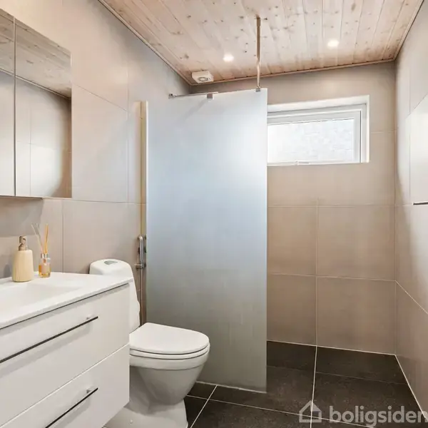 Badeværelse med hvid vask og toilet ved en frostet glasbruser, omgivet af beige fliser og et træloft. En lille vindue giver naturligt lys.