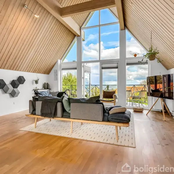 Stue med stor sofa centreret på et lyst tæppe. Store vinduer i loftshøjde viser himlen udenfor. Væggene har trækonstruktioner. Reoler med planter og tv står til højre.