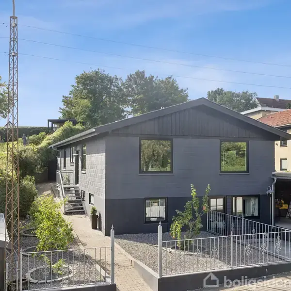 Et gråt hus med store vinduer står i en indhegnet have med grus. Der er indkørsel og en carport til højre. Omgivet af træer og nabohuse.
