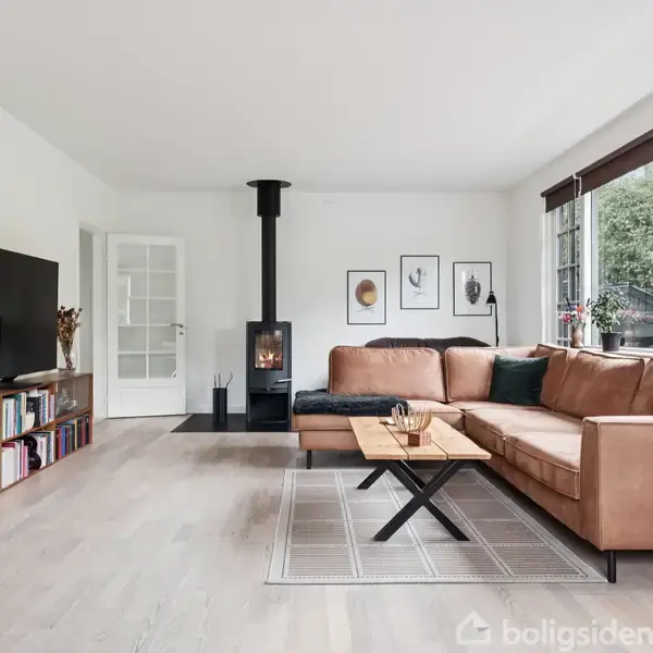 Brunt hjørnesofa står foran en sort brændeovn, omgivet af store vinduer med udsigt til haven. En fladskærms-TV er placeret på en reol fyldt med bøger.