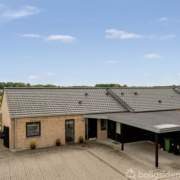 Rækkehus med fladt tag og mursten facade, parkeringsplads foran, beliggende i et åbent, landligt område med grønne marker i baggrunden og en klar blå himmel.