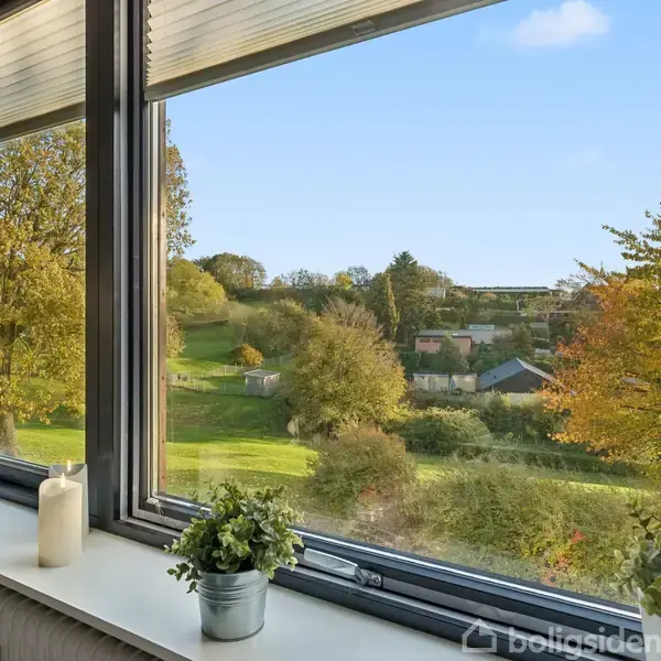 Vindue har udsigt til en grøn have med træer og buske under en blå himmel. Vindueskarmen har små planter i potter og et stearinlys. Udendørsområdet er frodigt og idyllisk.