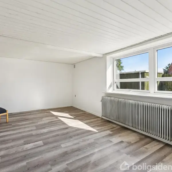 En enkel stol står stille i et lyst rum med trægulv og hvide vægge. Store vinduer og en radiator findes langs væggen, med udsigt til træbygninger udenfor.