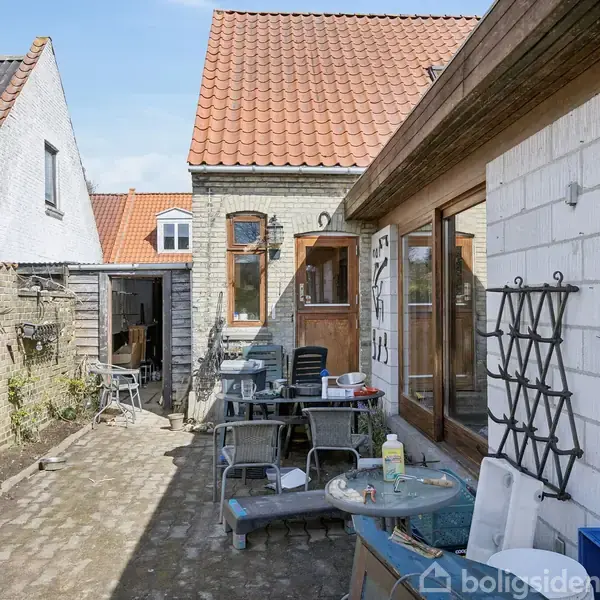 Hus med rødt tegltag og gul murstensfacade. Bord og stole står på den brostensbelagte gårdsplads. Hænger diverse dekorationer og værktøj på muren til venstre.