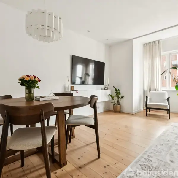Rundt spisebord med blomster står centralt. Området er lyst med træplankegulv, sofa og store vinduer. En stor plante ved vinduet. Vægmonteret TV.