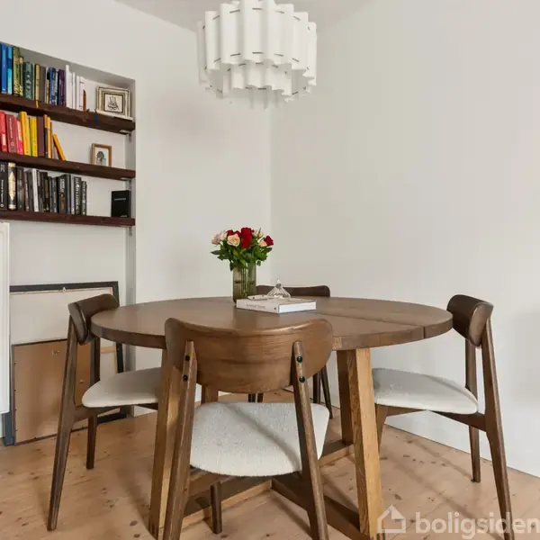 Rundt spisebord i træ med fire stole står i hjørnet af et lyst rum. På bordet står en vase med blomster. En bogreol står til venstre ved en radiator.