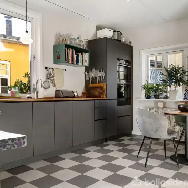 Et moderne køkken med grå skabe, en indbygget ovn og en bordplade ved et vindue. Grønne planter og krydderier er omhyggeligt arrangeret. Et lille bord og stol står ved siden.