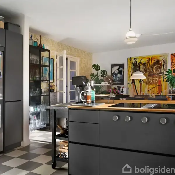 Grå køkkenø står centralt med kogeplader, omgivet af moderne skabe og farverig kunst på væggene. Skakternet gulv og hængende lamper skaber en moderne atmosfære.