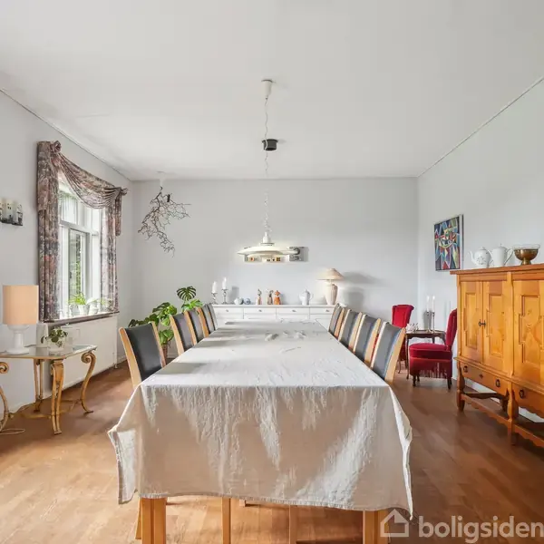 Langt spisebord med dug og stole i en spisestue. Rummet har træmøbler, lamper, og store vinduer med blomstrede gardiner, der lukker lys ind. En rød stol står ved væggen.