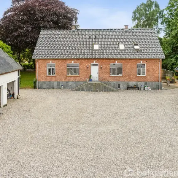 Rødt murstenshus med tegltag står i en gårdsplads omgivet af to hvidmalede lagerbygninger. Der er grusbelægning på jorden, og grønne træer omkranser området i baggrunden.