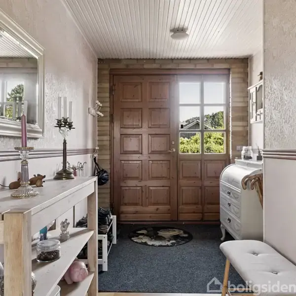 En entré med et trægulv indeholder et lille bord med dekorative genstande og et spejl til venstre. I midten er en stor trædør med små vinduer, der lukker naturligt lys ind.