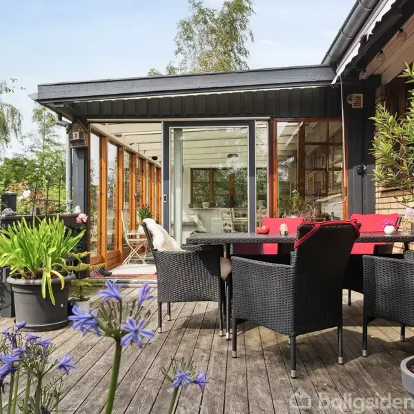 Terrassemøbler står på en træterrasse med plantekrukker, omkranset af et lyst haveområde. I baggrunden er der en bygning med store vinduespartier og en åben dør til indendørsområdet.