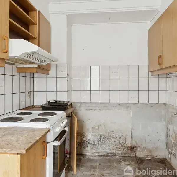 Komfur med fire kogeplader placeret i et køkkenhjørne, omgivet af brune overskabe, hvide fliser på væggen og tomme hylder. Køkkenets gulv og vægge viser tegn på slid.