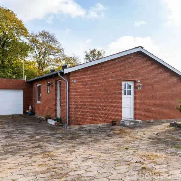 En enkel, rød murstensbungalow med en hvid dør og tilstødende garage. Ejendommen er omgivet af en flisebelagt indkørsel og omkredset af grønne træer under en blå himmel.