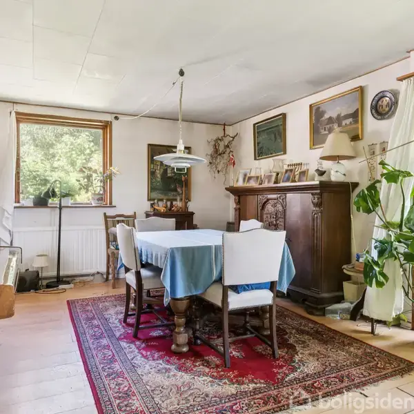 Et spisebord med blå dug er omgivet af træstole i en stue, hvor der også er et klaver til venstre. Rummet har grønne planter, billeder på væggene og store vinduer.