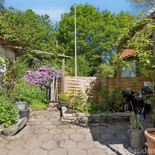 En have med fliser omgivet af grønne planter og træer. En flagstang står centralt, og et træhegn skaber en privat atmosfære. Planter i potter er placeret langs flisekanten.