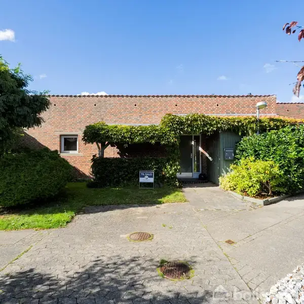 Murstenshus med grøn bevoksning omkring dørkarmen. Huset står i en lille have med græs, træer og en flisebelagt sti i et solrigt område.