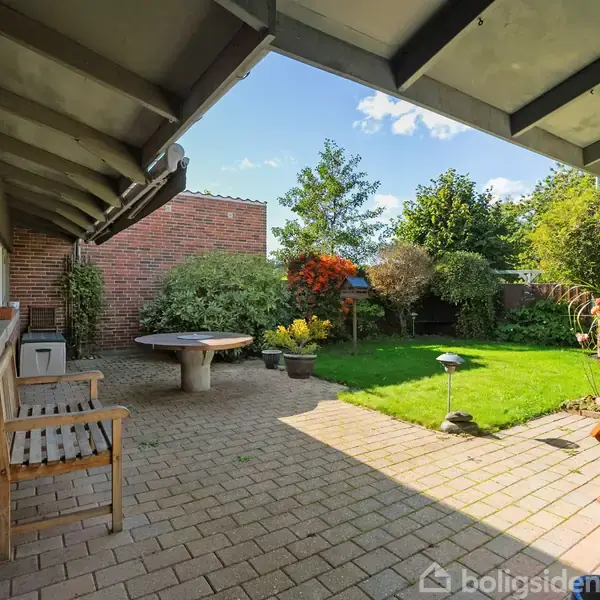 Terrasseområde med træbænk og bord står stille. Omgivet af en have med frodige buske og træer, under en halvoverdækket struktur, som giver skygge på solrige dage.