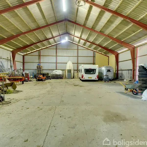 En rummelig lade med et højt loft, indeholdende campingvogne og diverse landbrugsudstyr på gulvet. Vægge og tag har en metalstruktur. Omgivelser præget af opbevaring og orden.