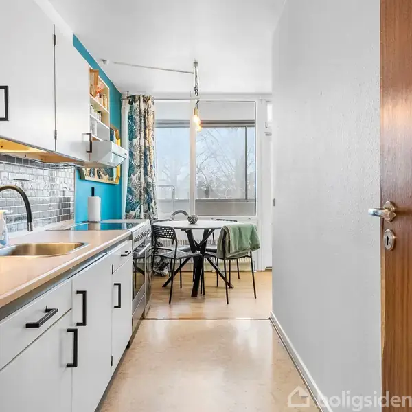 Køkken med hvidt køkkenskab og vask til venstre, pyntet køleskabsdør. Spisebord med stole foran stort vindue i baggrunden, lysektrært hængende fra loftet, gardin ved siden af vinduet.