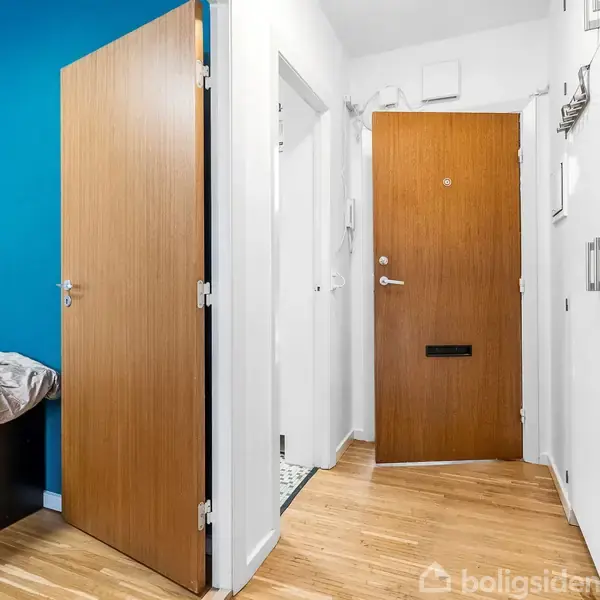 En gang med trægulv og hvidmalede vægge. To åbne døre fører ind til forskellige rum. Der er en blåmalet væg til venstre med en seng synlig.