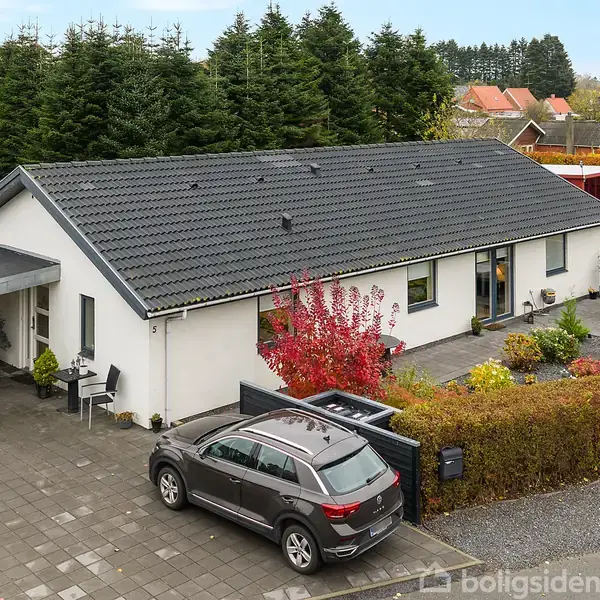 En moderne bungalow med sorte tegltag og hvide vægge står ved en indkørsel. En grå SUV er parkeret foran huset. Omgivet af velplejede hække og træer i villakvarter.