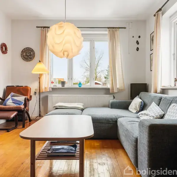Grå sofa i L-form står i lys stue, belyst af moderne loftlampe. Stuen har brune trægulve, et stort vindue med udsigt og flere små pyntegenstande.