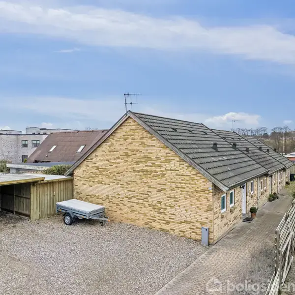 En række gulstensrækkehuse står langs en flisebelagt sti i et boligområde. En bil er parkeret under en carport, og en trailer er placeret på gruset ved siden af.