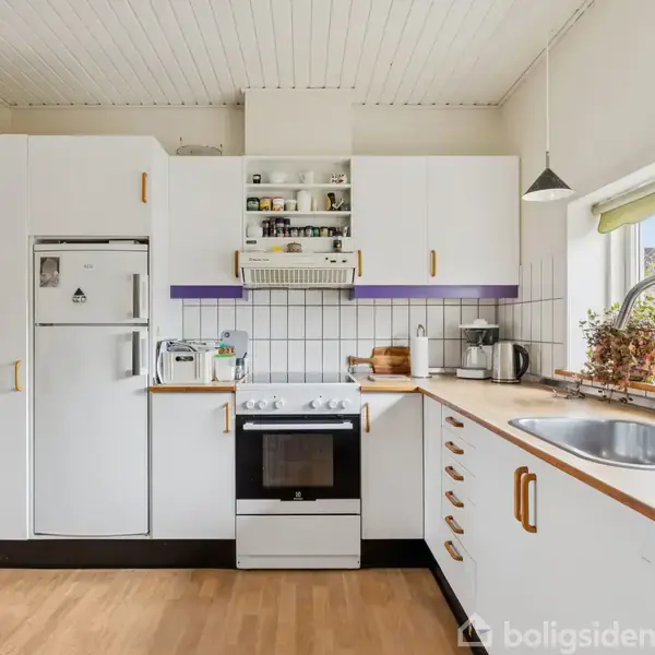 En lys køkkenindretning med hvidt køleskab og komfur, omgivet af hvide skabe og træbordplade. Et vindue med grønne planter ved siden af vasken tillader naturligt lys.