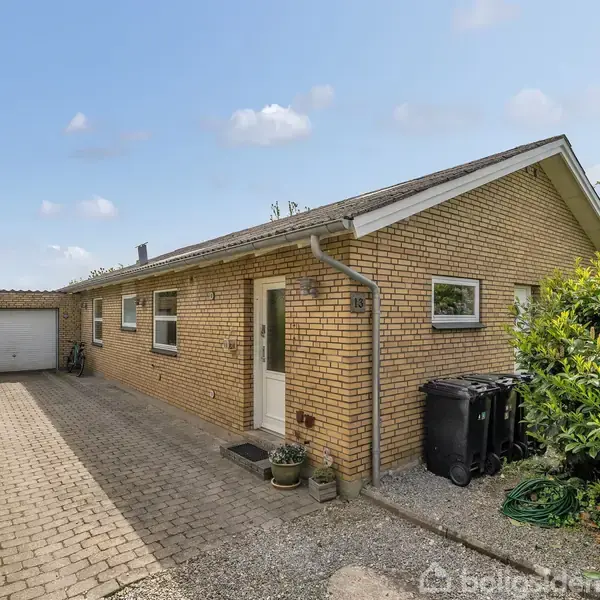 Et gult murstenshus med et enkelt etage står stille ved siden af en flisebelagt indkørsel. En garage ses i slutningen af indkørslen, omgivet af grønne buske og blå himmel.