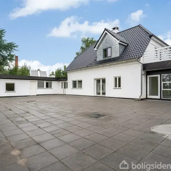 Hvidt hus står stille ved en stor, flisebelagt terrasse. Omkring huset er der træer og grønt, mens himlen er blå med få skyer.