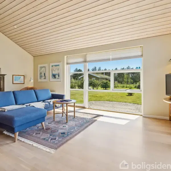 Blå sofa står i et lyst rum med trægulv, vendt mod et stort vindue med udsigt til en have. En reol med tv står til højre.