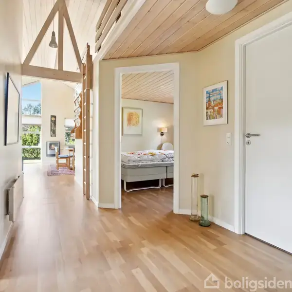 En gang med trægulv, lysindfald fra store vinduer og åbne døre ind til et soveværelse. Rummene er lyse med moderne indretning og træloft. Udsigt til en have gennem vinduerne.