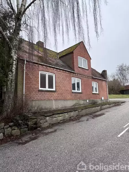 Rød murstenshus med grøn mos på taget står ved en vej. Omkringliggende træer og grå himmel skaber en vinterlig atmosfære.