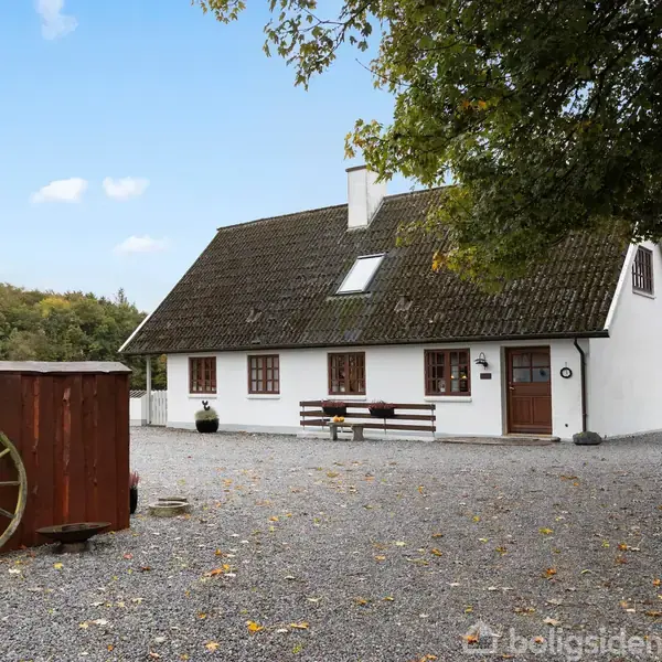 Huset står stille i en gårdsplads med grus, omgivet af træer og marker. Det har hvid facade og mørkt tag. I forgrunden ses en trævogn med et stort hjul.