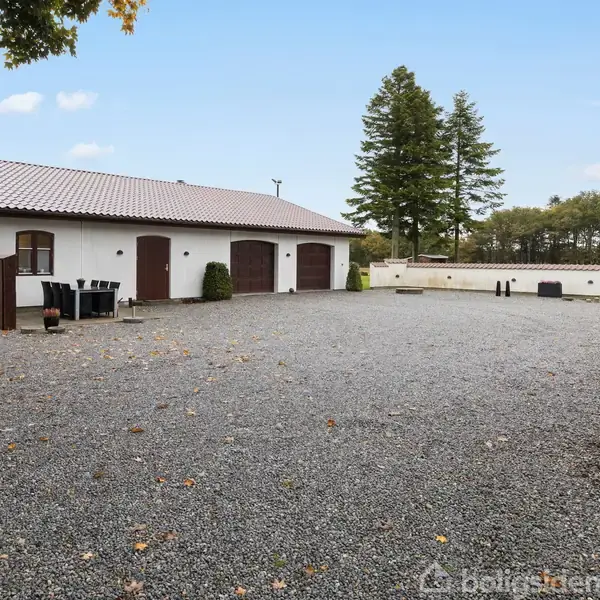 Grusbelagt gårdsplads med en hvid bygning med brun tag og tre porte til venstre. To caféborde med stole står udenfor. Træer og grønne marker i baggrunden.