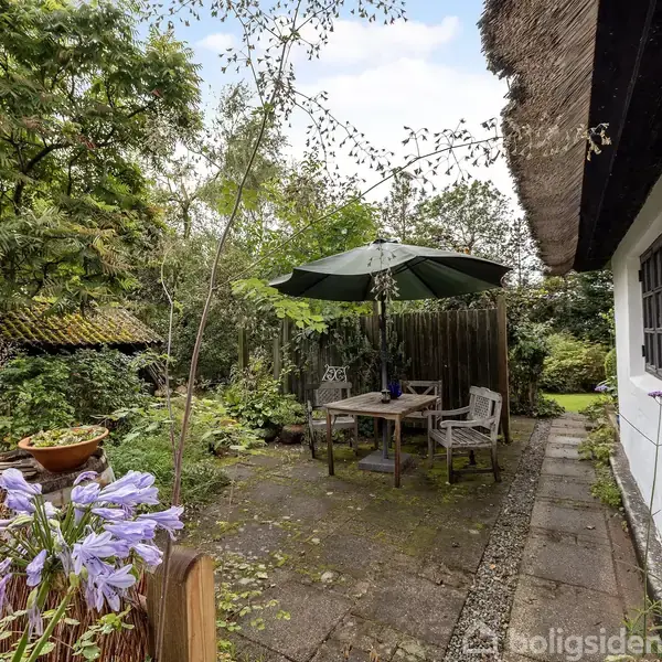 Et bord med stole står under en parasol på en stenbelagt terrasse ved siden af et hvidt hus med stråtag. Omgivet af blomstrende planter og træer.
