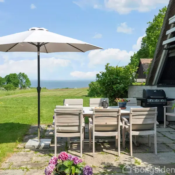 En terrasse med et havebord og stole står under en parasol. En grill er placeret ved et hus i nærheden. Omgivelserne er en grøn have med udsigt til havet.