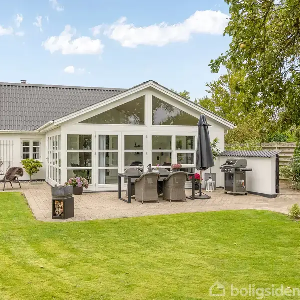 Et hvidt hus med store vinduespartier vender ud mod en terrasse med spisebord og parasol. Det er omgivet af en velplejet græsplæne og træer.