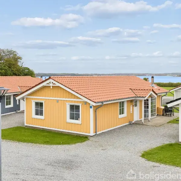 Gul træhytte står stille mellem grusveje, flankeret af røde og blå hytter. Baggrunden viser vand og skov, himlen er let skyet.