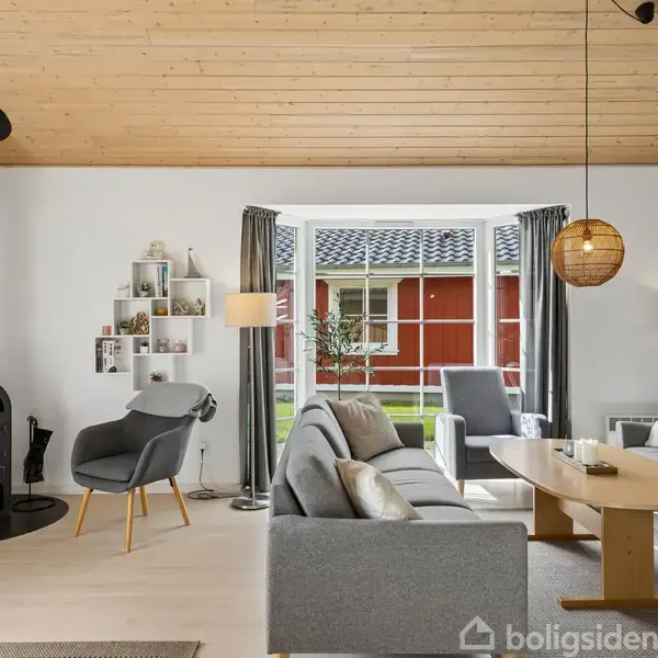 Pejs står ved væggen, brænder stille i en stue med lyse træmøbler. Stuen har en stor vindue, der viser en rød bygning udenfor. Gråt sofa-område skaber hyggelig atmosfære.
