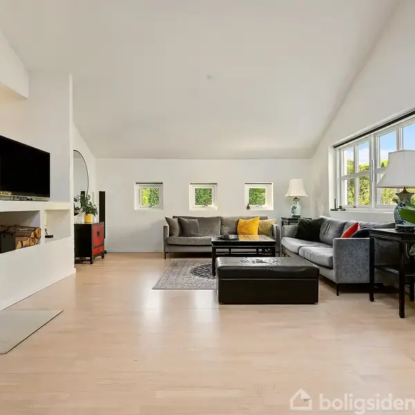 En lys stue med en grå sofagruppe, sofabord og en pejs. Et stort vindue på højre side giver udsigt til grønne træer. Planter står ved siden af sofaen.