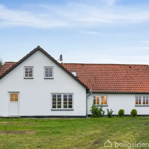 Et hvidt hus med rødt tegltag står på en grøn græsplæne. Huset har vinduer med sprosser og en dør. Baggrund består af træer og en blå himmel.