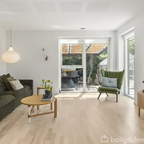 En stue med en grøn sofa og stol (objekt) står ved siden af et rundt sofabord (handling). Store vinduer i baggrunden giver udsigt til en terrasse (kontekst).