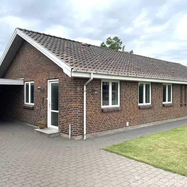 En murstensbungalow med rødt tag står stille. Omgivet af en flisebelagt indkørsel og velholdt græsplæne, viser huset hvide trærammer omkring vinduer og en glasdør nær en enkelt plantepotte.