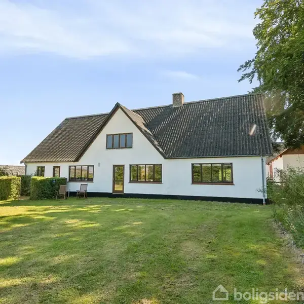 Hvidt hus med sort tag står stille i en have med velplejet græsplæne. Omgivet af buske og træer under en blå himmel.