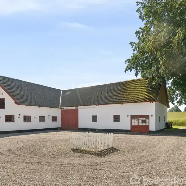 Gårdbygninger med hvid facade og mørkt tag omkranser en grusgård med en lille hvid indhegning. Træ og mark ses i baggrunden. "1959" står skrevet på bygningen.